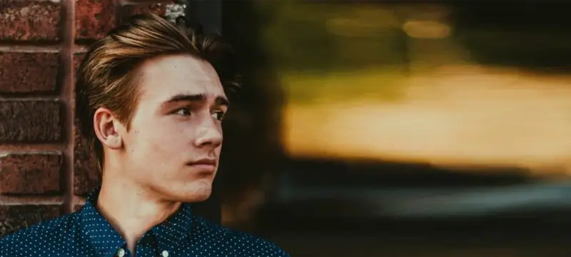 Junger Mann mit stylischen herren frisuren mittellang, lehnt an einer Ziegelwand.